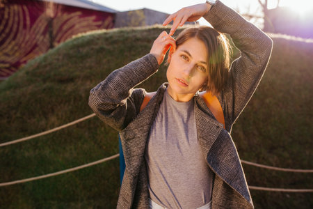 Young beautiful woman with short hair in a light grey fashionable coat with a backpack is posing for...