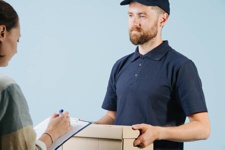 Woman is accepting a box delivery from a courier by signing paper form with a pen. Cropped. Delivery man is dressed in dark blue uniform.の写真素材