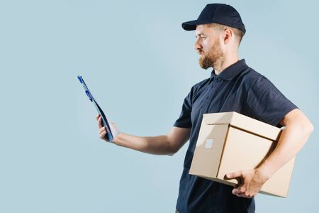 Delivery man with serious face is holding his orders list over blue background. He is checking it while holding box under arm. He dressed in a dark blue uniform. Side view.の写真素材