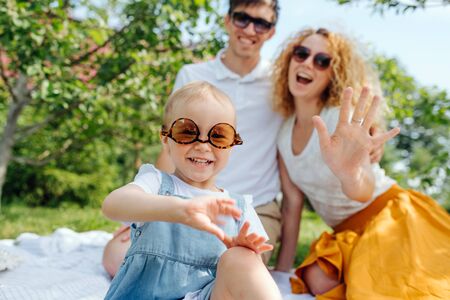 Summer family picnic on grass in the gardens. Mother, father and their precious infant daughter, all in sunglasses, are sitting on white tablecloth. Laughing together, posing for a photo.の写真素材