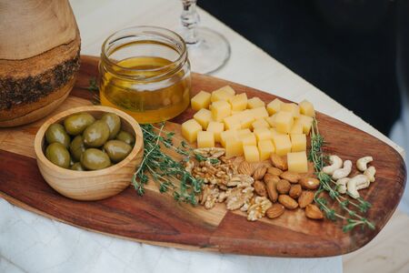 Variety of appetizers for wine such as: olives, cheese, honey, nuts, thyme, served on a wooden plate of a special form. All on a crude plank improvised table, covered with white tablecloth.の写真素材