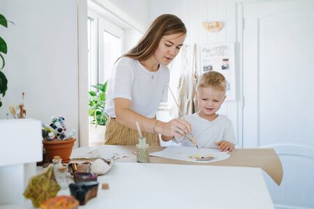 Young mother and first grade son are painting together at home. Lots of painting supplies lie on table next to them. Light walls, bright room.の写真素材