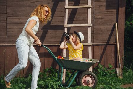 Happy mother driving a wheelbarrow fast, while her teenage daughter riding in it, looking at her through camera, taking picture. Summer recreation in the gardens next to a country house.の写真素材