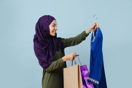 young smiling muslim girl in casual clothes with shopping bags chooses a party dress. Studio shot. Isolated blue backgroundの写真素材