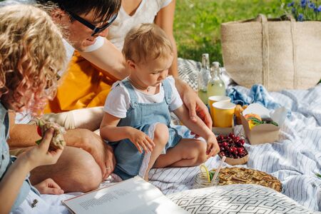 Summer family picnic on grass in gardens under shade of trees. Mother, father, two children sitting on white tablecloth next to their food. Being attentive to small one. Dessert is coming up soon.の写真素材