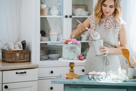 Caucasian blond woman wearing an apron whips sweet curd cream for biscuit cake, standing at home...