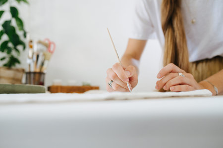Young woman artist with unfixed braid is painting on sheet, behind the table at home. Cropped, no...