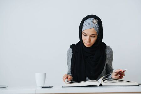 young cute muslim student sitting at the table and reading a book. Studio shot. Isolated white backgroundの写真素材