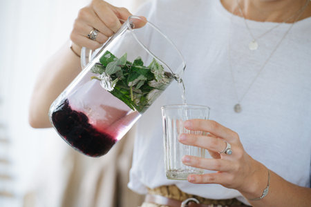 Young woman is pouring fresh lemonade made from berries and mint from jug into glass at home. She is...