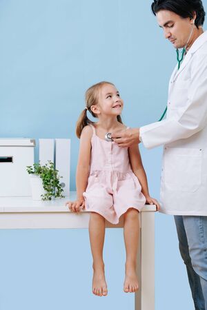 Male doctor examining a cheerful first grader child girl with two tails by using a stethoscope. She sits on a table in a hospital, her legs hanging. Blue wall, white objects.の写真素材