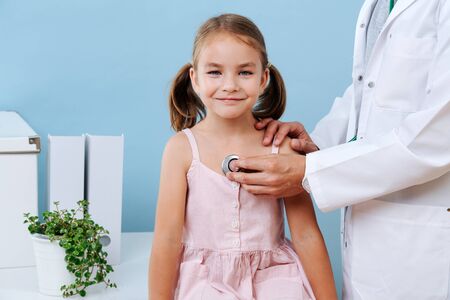 Male doctor hands examining a cheerful first grader child girl with two tails by using a stethoscope. Listening to her heartbeat and breathing. Blue wall, white objects. She looks straight at camera.の写真素材