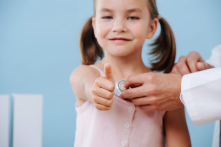 Male doctor hands examining cheerful first grader child girl with two tails by using a stethoscope. Listening to her heartbeat. Blue wall, white objects. She looks straight at camera, shows thumbs up.の写真素材