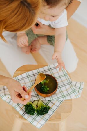 Active jolly infant child eating broccoli soup. Sitting on his mother's knees.の写真素材