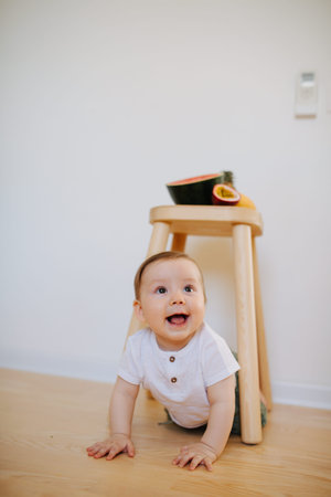 An infant brunet baby boy is crawling from under the stool with fruits on it