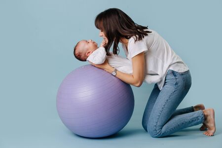 Happy mother carefully and gently doing exercises with her infant child baby on purple yoga ball over blue background. Baby is lying on the back, while they play Their eyes are clothed.の写真素材