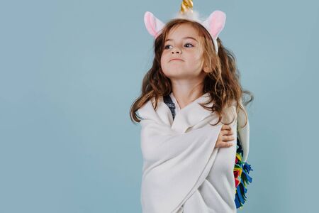 Adorable little preschooler girl with brown wavy hair is standing in noble stance, wearing unicorn headband, wrapped in bed cover over blue background. Engaged in imaginative role play.の写真素材
