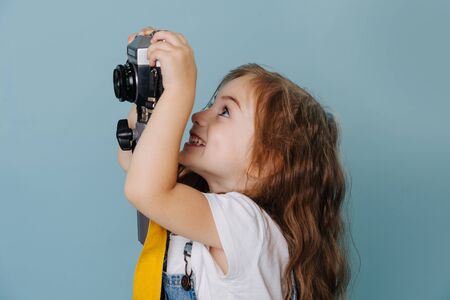 Little preschooler girl with brown wavy hair is trying to take an image with a vintage mirrored camera, fixed high on a tripod. Over blue background. Side view.の写真素材