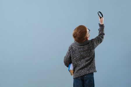 Portrait of a cute smiling little ginger boy in glasses looking up through a magnifying glass over blue background. He's wearing grey buttoned hooded sweater. Half length. From behind.の写真素材