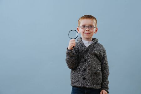 Portrait of a cute smiling little ginger boy in glasses with magnifying glass in his hand over blue background. He looks pleased with himself. He's wearing grey buttoned hooded sweater. Half length.の写真素材