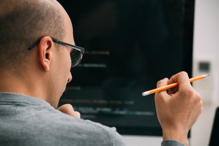 Bald caucasian programmer in glasses is sitting behind the desk, in front of black monitor, looking...