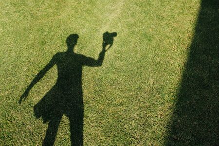 The shadow of the photographer with camera on the freshly mowed green lawn at mid summer sunny day. He has a bag on his side, his left hand slightly extended.の写真素材