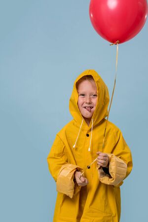 Little boy in yellow raincoat with red balloon over blue background. Horror movie style for halloween. With grimace and tongue out. Half length.の写真素材