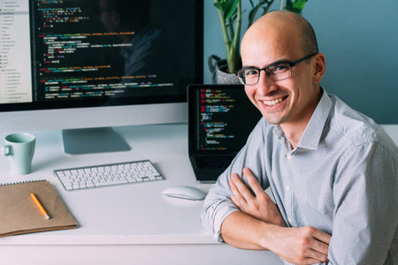 Bald caucasian programmer in glasses is sitting behind the desk, in front of two black screens,...