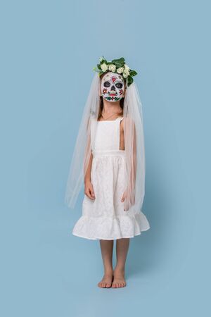 Little girl in bridal gown, veil and hair wreath wears painted scary halloween skull mask over blue background. She stands still and unalive, looks at camera. low angle.の写真素材