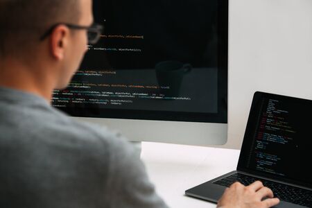 Bald caucasian programmer in glasses is sitting behind the desk, in front of two black screens, laptop and monitor, looking closely, analysing code lines. He's very attentive and focused.の写真素材