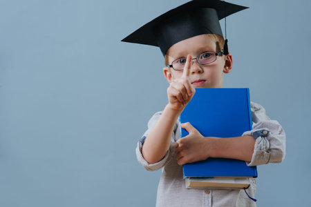 Cute smiling little ginger boy in glasses, wearing student hat holds stack of books in hands over...
