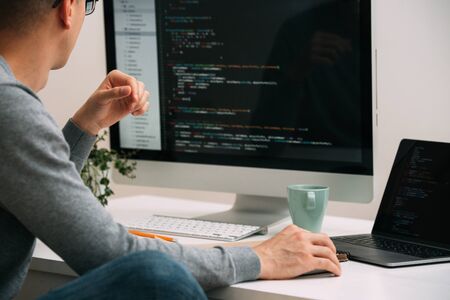Bald caucasian programmer in glasses is sitting behind the desk, in front of two black screens, laptop and monitor, looking closely, analysing code lines. He's very attentive and focused.の写真素材