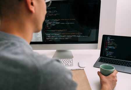 Bald caucasian programmer in glasses is sitting behind the desk, in front of two black screens, laptop and monitor, analysing code lines. He's holding onto a green mug.の写真素材