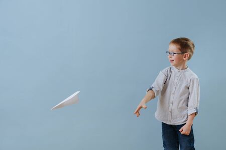 Cute little ginger boy launched a paper plane to fly over blue background. He's very happy about the experience. Side view.の写真素材