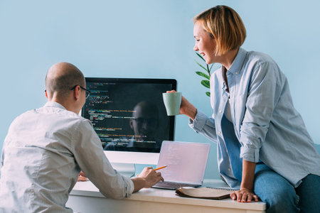 Bald caucasian programmer in glasses is sitting behind the desk, in front of black screen, filled...