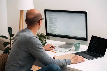 Bald caucasian programmer in glasses is sitting behind the desk, in front of two screens, laptop and monitor, one black, full code lines, other white with google search.の写真素材