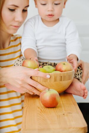 Jolly blond toddler boy is sitting on high narrow table. Leg at each side. He explores, plays with apples from backet. Touching them. Mother watches so backet wouldn't fall.の写真素材