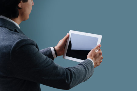 Businessman in a gray suit is holding a tablet with both hands, looking on a black screen over blue...