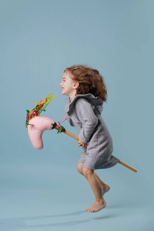 Little preschooler girl with brown wavy hair is hopping on a hobbyhorse toy, hovering in the air over blue background. Indoors, side view.の写真素材