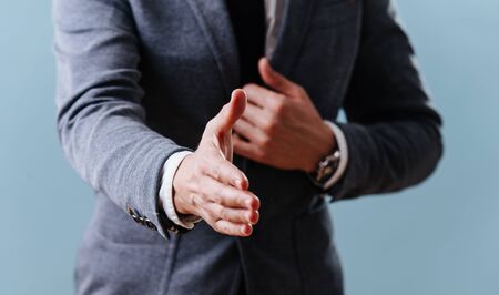 Businessman in a gray suit is extending his right hand to do a handshake, holding lapel with his second hand over blue background. Cropped, no head.の写真素材
