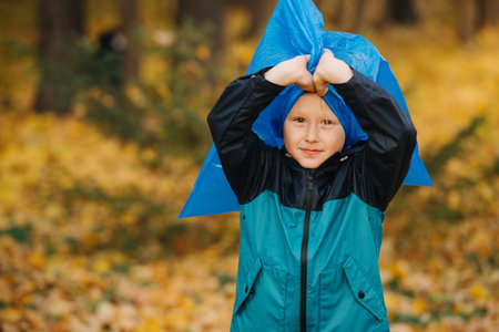 Little first grader child with a trash bag in a seasonal forest at autumn, helping to clean it with a group of volunteers. He's playing with a bag, wearing it like a headwearの写真素材