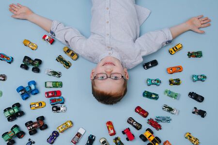 Happy little ginger boy lying on the floor surrounded by toy model cars all around him. They placed in half-circular shape in several rows. Over blue background. He extended hands showing his treasureの写真素材