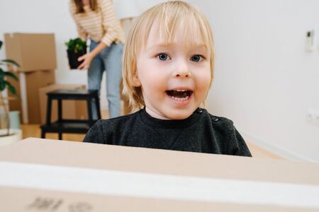 Woman packing. Her little son carries boxes and arranges them in a stack. Moving out, relocating. Boy is having fun, running along with box in hands, being enthusiastic about his experience. Close up.の写真素材