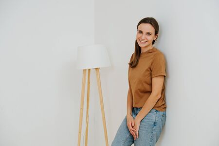 Smiling woman, leaning her back against the wall, next to a floor lamp in corner. She's of middle age, casually dressed, wears long brown hair. Half length.の写真素材