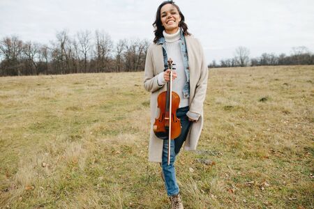 Happy woman in a coat laughing as she walks through a field of short dry grass with violin in hands. She's in the middle of a clearing in a forest of leafless autumn trees.の写真素材