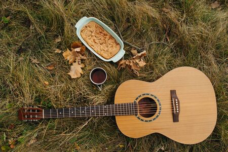 Six strings classic guitar lying on an autumn green and dry yellow grass next to a cup of tea and a pie in a form. Top view.の写真素材