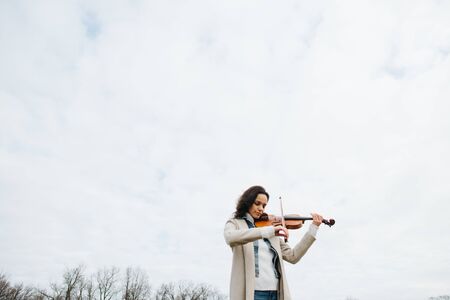Beautiful woman in a coat playing violin under a sky with her eyes closedの写真素材