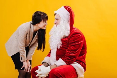 Young Santa claus with lowered beard chatting with attractive brunette. She leaned forward, very close to his face. They are looking into each other's eyes and giggling. Over yellow background.の写真素材