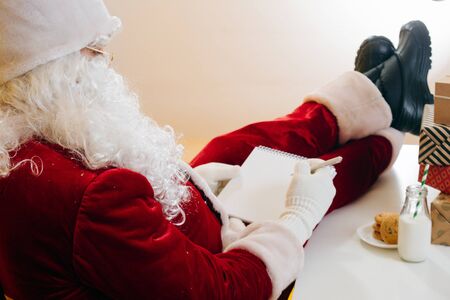 Santa Claus leaning on the back of a chair, put his legs on the table next to a pile of presents. He's composing his orders list. Over yellow background. From behindの写真素材