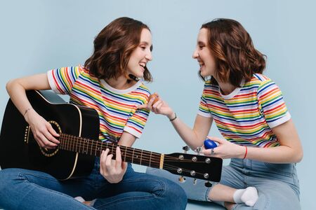 Portrait of two twin sisters in colored rainbow t-shirts sing and play on guitar and percussion togetherの写真素材