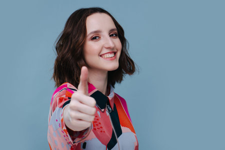 Happy girl holding forward hand and raising thumb up, wearing white t-shirt isolated blue color backgroundの写真素材